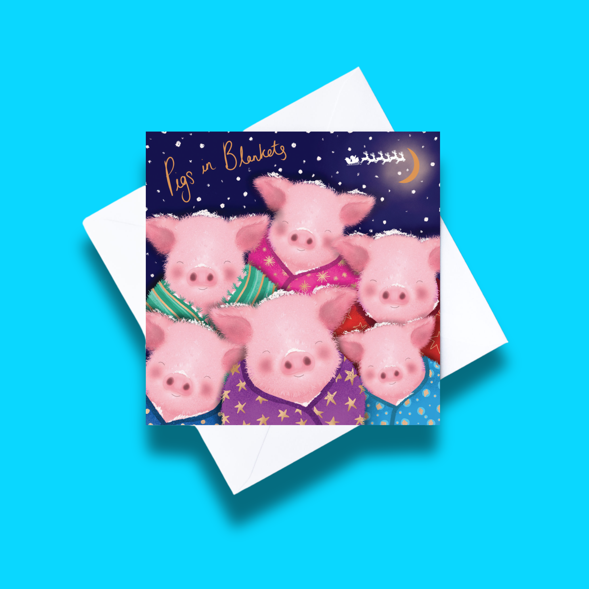 Pigs in Blankets Christmas card with white envelope on a blue background. Card design features 6 pigs in blankets on a dark starry background and text that says pigs in blankets