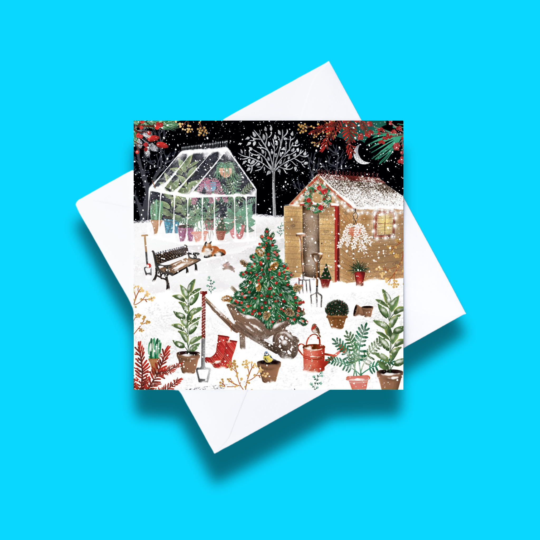 Christmas Garden Christmas card with white envelope on a blue background. Card design features a snowy garden during Christmas time with a shed, greenhouse and various plants